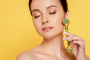 beautiful naked woman with closed eyes using jade roller on face isolated on yellow