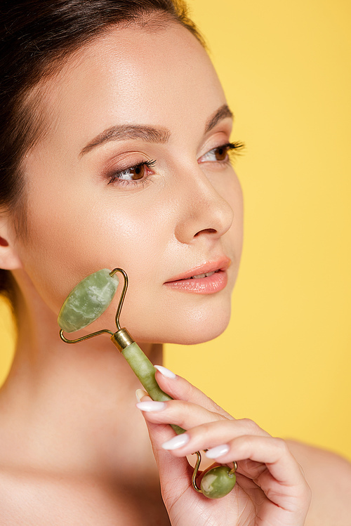 portrait of beautiful naked woman using jade roller on face isolated on yellow