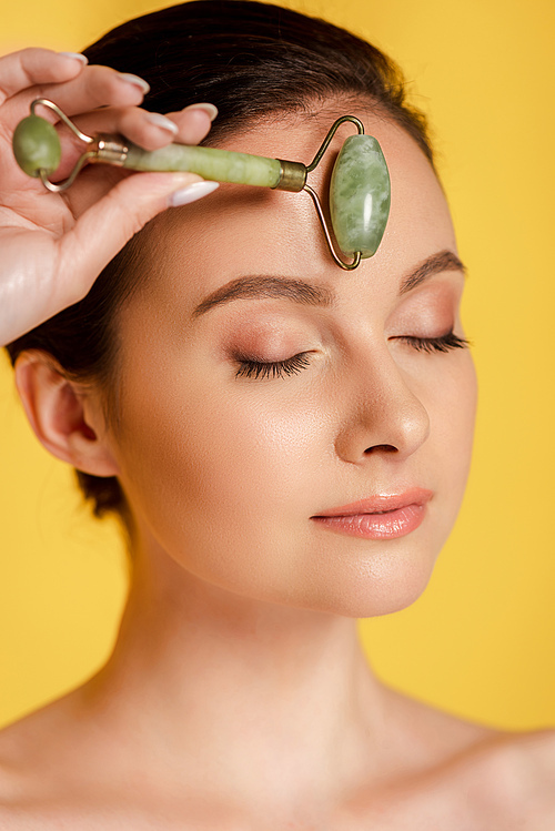 beautiful naked woman with closed eyes using jade roller on face isolated on yellow