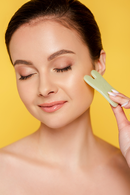 portrait of beautiful naked woman with closed eyes facial jade gua sha isolated on yellow