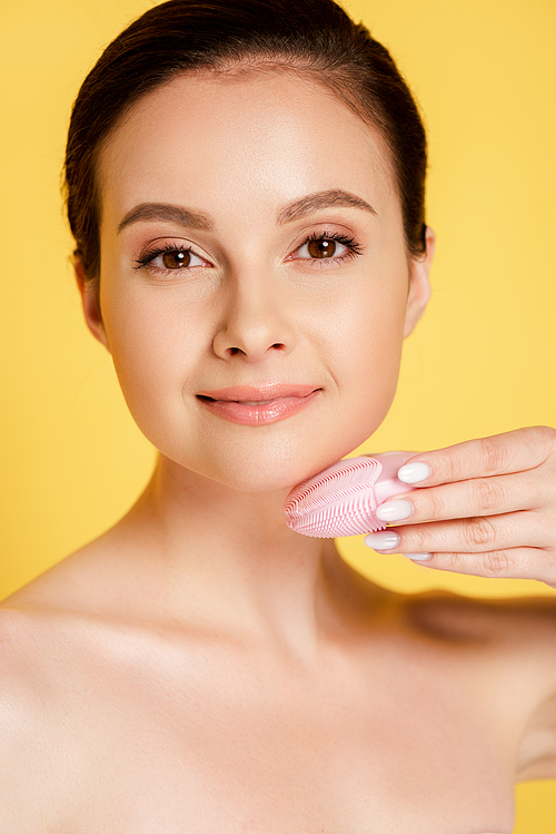 beautiful naked woman using facial cleansing brush isolated on yellow