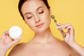 naked beautiful woman holding cosmetic cream and jade roller isolated on yellow