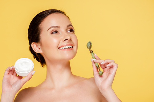 smiling naked beautiful woman holding cosmetic cream and jade roller isolated on yellow