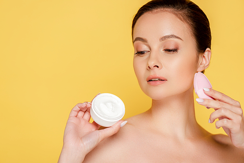 naked beautiful woman holding cosmetic cream and facial cleansing brush isolated on yellow