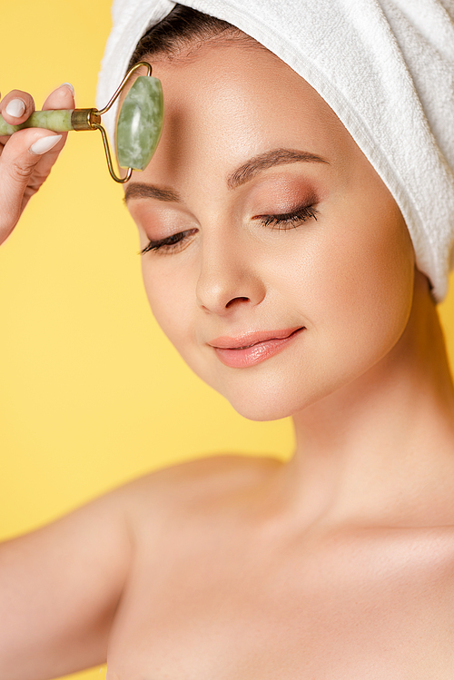 beautiful naked woman with towel on head using jade roller isolated on yellow