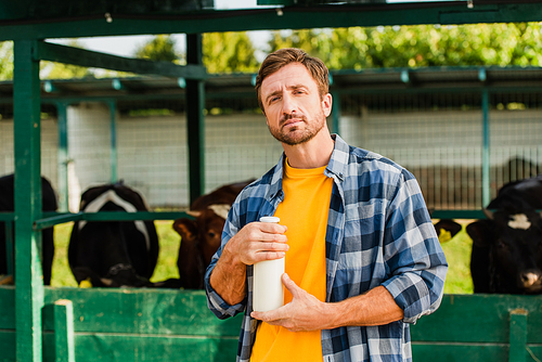 rancher in checkered shirt holding bottle of fresh milk while  near cowshed