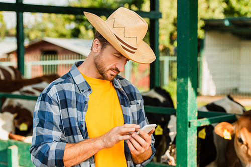farmer in checkered shirt and straw hat chatting on cellphone on farm near cowshed