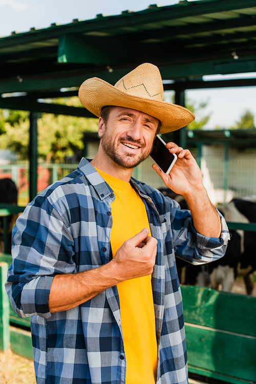 rancher in straw hat and plaid shirt talking on smartphone near cowshed