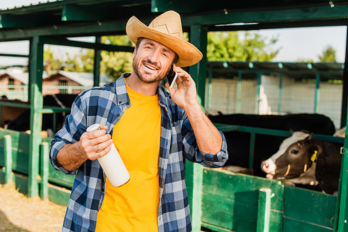 farmer in checkered shirt and straw hat holding bottle of fresh milk while talking on smartphone