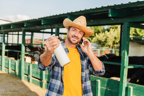 rancher in straw hat and plaid shirt talking on mobile phone while showing bottle of fresh milk near cowshed