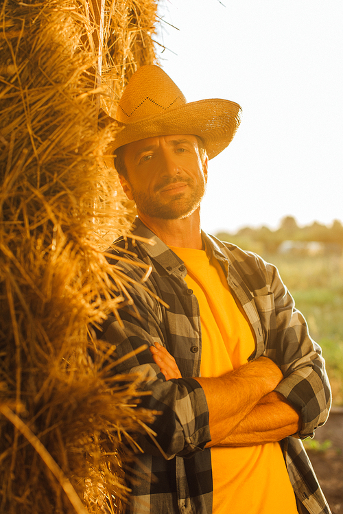 rancher in checkered shirt and straw hat  while leaning on bale of hay with crossed arms