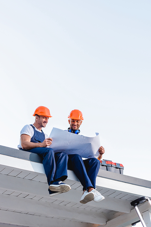 Low angle view of workmen holding blueprint while sitting near toolbox on roof of building