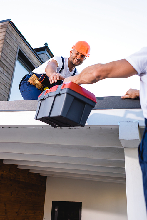 Selective focus of builder giving toolbox to colleague on roof of house