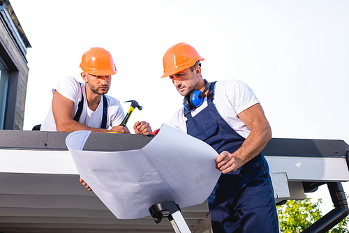 Handyman holding blueprint while standing on ladder near colleague with hammer on roof of building