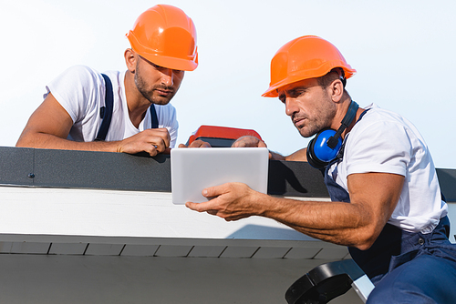 Selective focus of handymen in workwear using digital tablet while working on roof of building