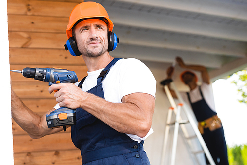 Selective focus of handyman in workwear and ear defenders holding electric screwdriver near facade of building