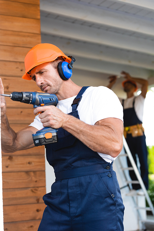 Selective focus of manual worker in helmet and ear defenders holding electric screwdriver near facade of house