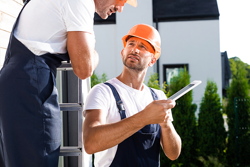 Selective focus of builder in overalls holding digital tablet near colleague on ladder and house