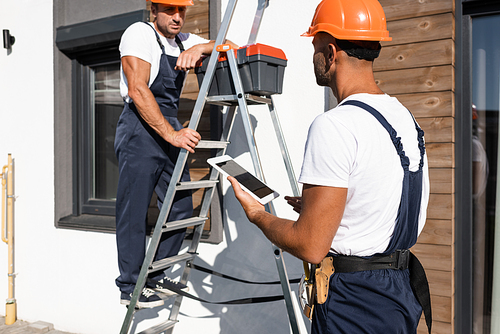 Selective focus of handyman with digital tablet standing beside colleague in uniform on ladder and house
