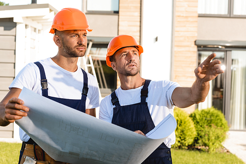 Builder pointing with hand near colleague with blueprint outdoors