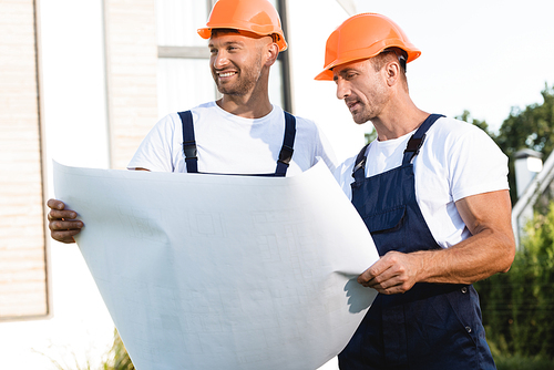 Builders in hardhats and uniform holding blueprint outdoors