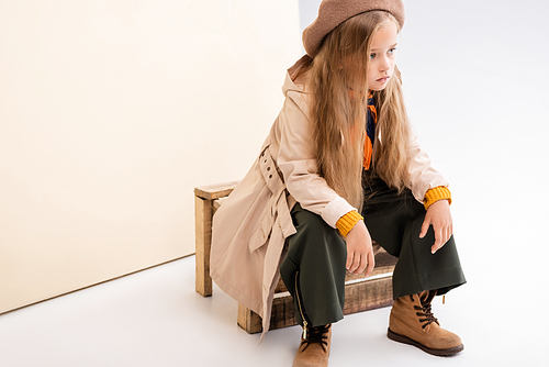 fashionable blonde girl in autumn outfit sitting on wooden box on beige and white background