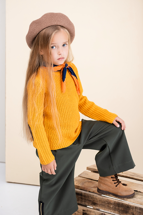 fashionable blonde girl in autumn outfit posing on wooden box on beige and white background
