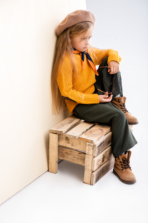 fashionable blonde girl in autumn outfit with sunglasses sitting on wooden box on beige and white background