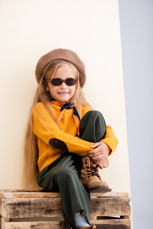 fashionable blonde girl in autumn outfit and sunglasses sitting on wooden box on beige and white background