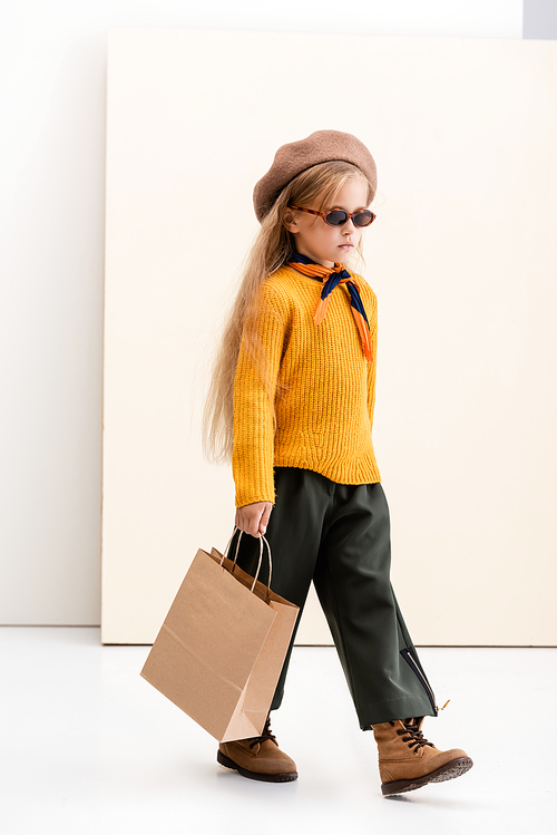 fashionable blonde girl in autumn outfit and sunglasses walking with shopping bag on beige and white background
