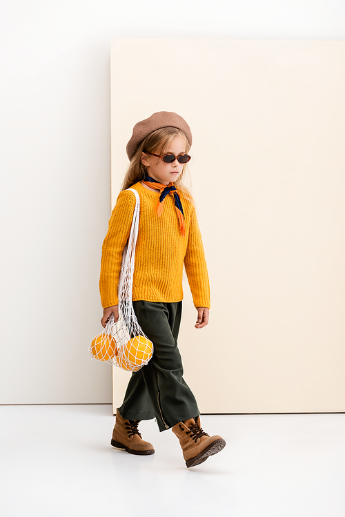 fashionable blonde girl in autumn outfit and sunglasses walking with grapefruits in string bag on beige and white background