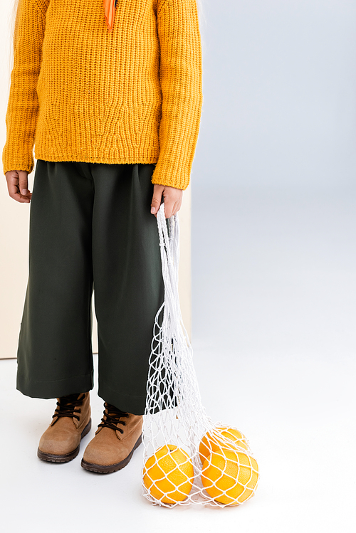 cropped view of fashionable girl in autumn outfit posing with grapefruits in string bag on beige and white background