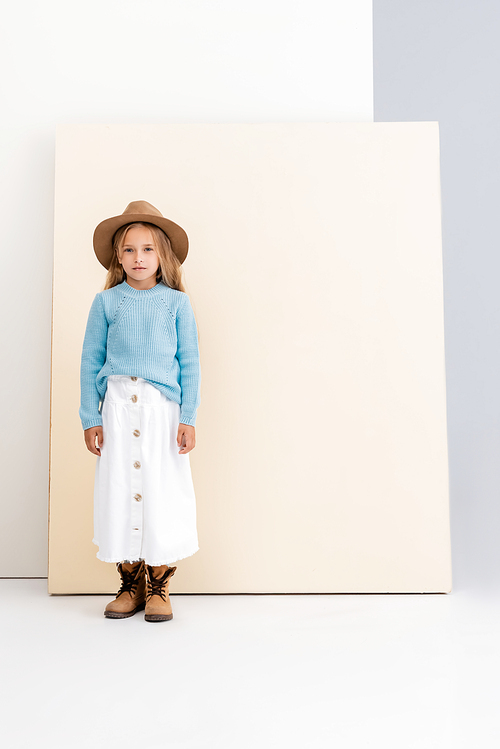 fashionable blonde girl in brown hat and boots, white skirt and blue sweater near beige wall