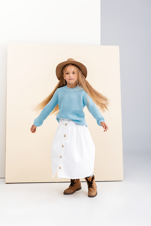 fashionable blonde girl in brown hat and boots, white skirt and blue sweater spinning near beige wall