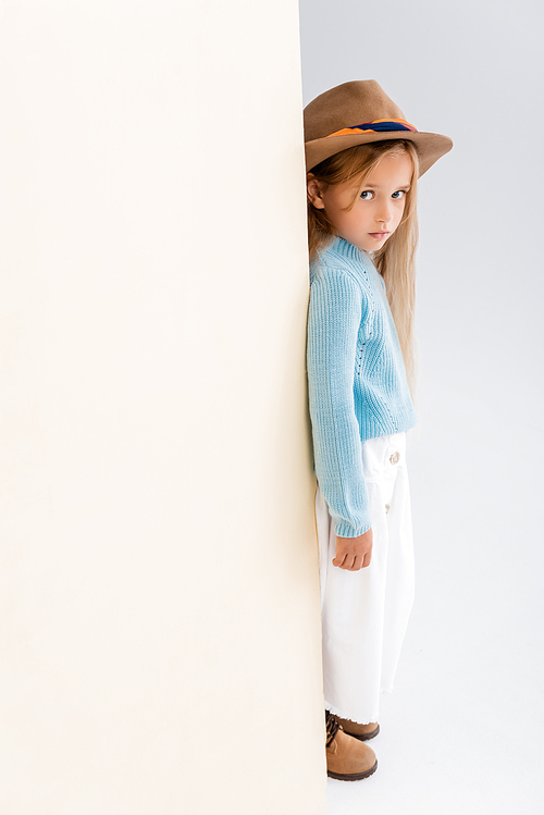 side view of fashionable blonde girl in brown hat and boots, white skirt and blue sweater near beige wall
