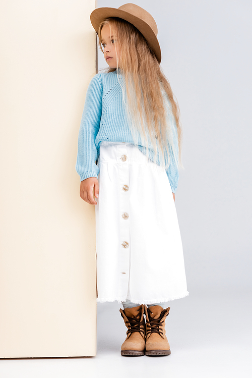 fashionable blonde girl in brown hat and boots, white skirt and blue sweater near beige wall