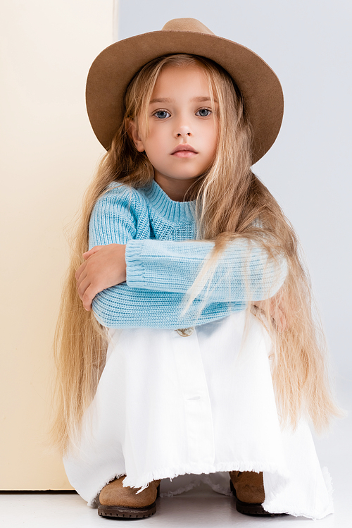 fashionable blonde girl in brown hat and boots, white skirt and blue sweater sitting near beige wall