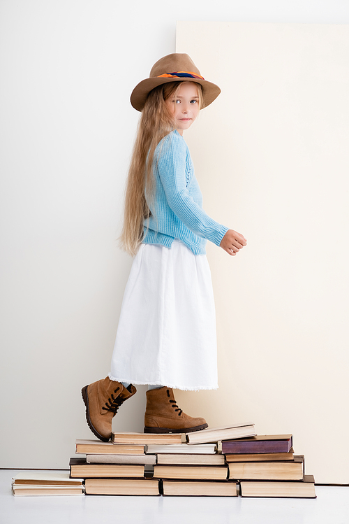 side view of fashionable blonde girl in brown hat and boots, white skirt and blue sweater walking on vintage books