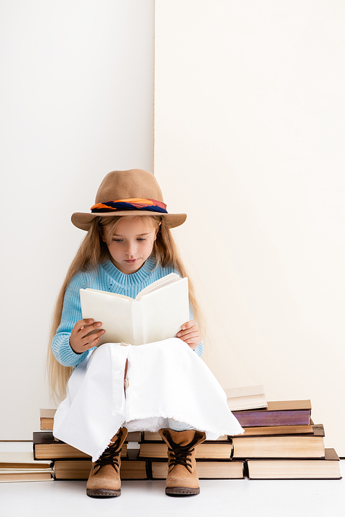 fashionable blonde girl in brown hat and boots, white skirt and blue sweater sitting on vintage books and reading near beige wall