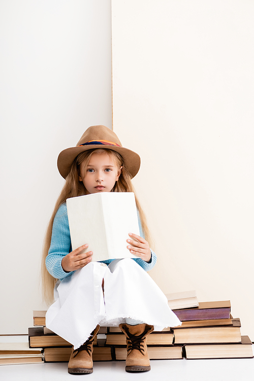 fashionable blonde girl in brown hat and boots, white skirt and blue sweater sitting with book near beige wall