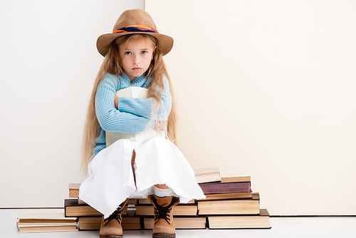 sad fashionable blonde girl in brown hat and boots, white skirt and blue sweater sitting with book near beige wall