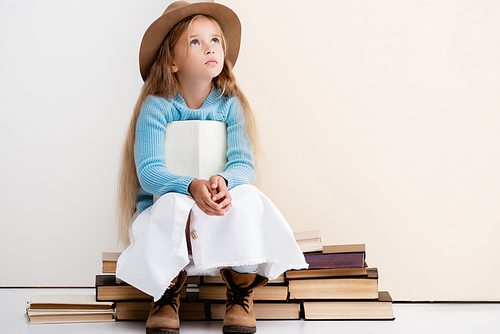 dreamy fashionable blonde girl in brown hat and boots, white skirt and blue sweater sitting with book near beige wall