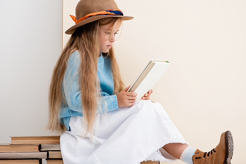 fashionable blonde girl in brown hat and boots, white skirt and blue sweater sitting on vintage books and reading near beige wall