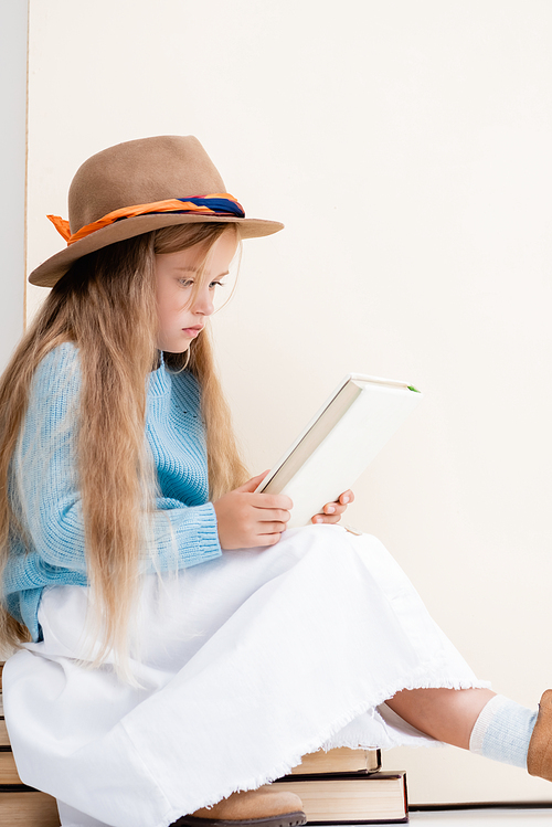 fashionable blonde girl in brown hat, white skirt and blue sweater sitting on vintage books and reading near beige wall