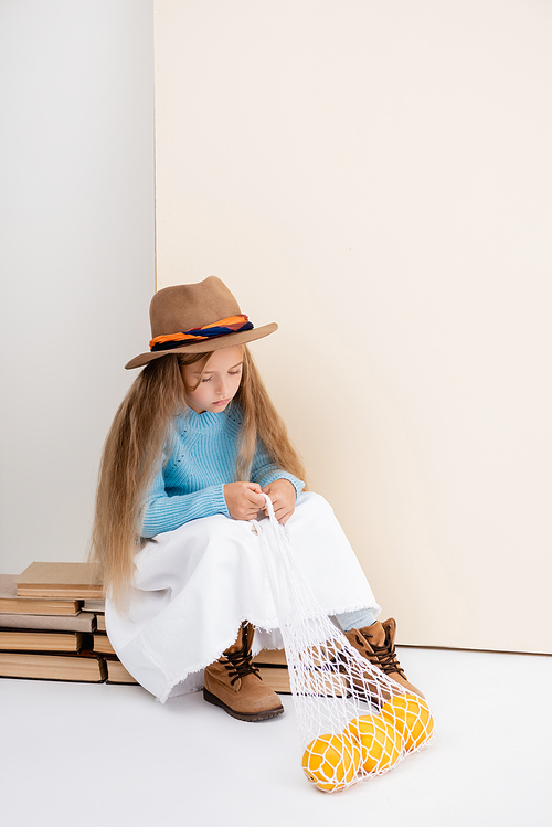 fashionable blonde girl in brown hat and boots, white skirt and blue sweater sitting on vintage books with grapefruits in string bag