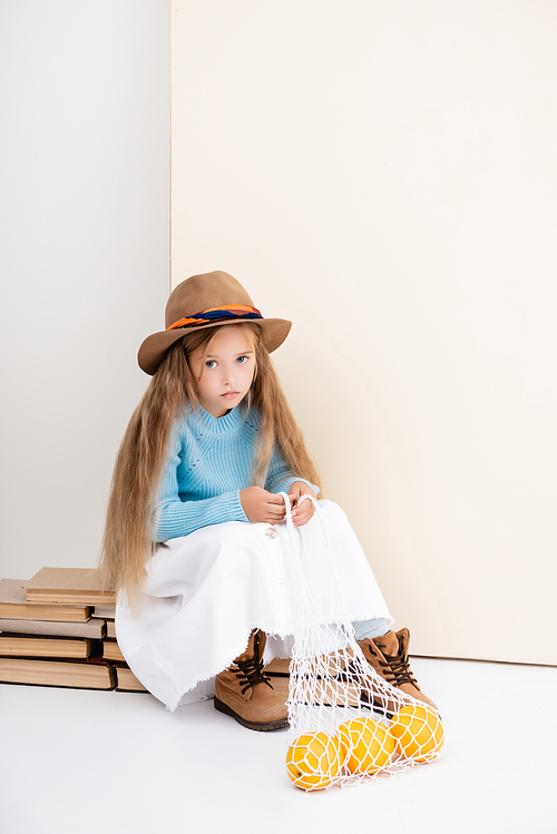 fashionable blonde girl in brown hat and boots, white skirt and blue sweater sitting on vintage books with grapefruits in string bag