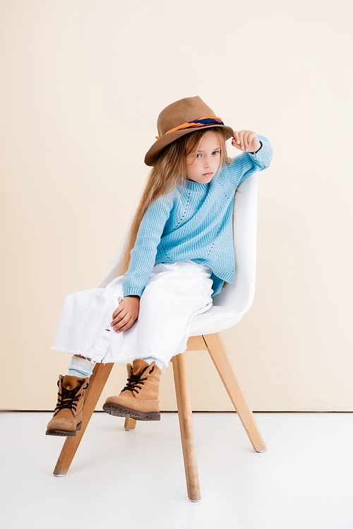 fashionable blonde girl in brown hat and boots, white skirt and blue sweater sitting on chair near beige wall