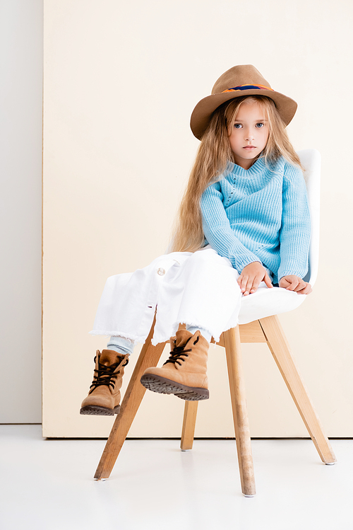 fashionable blonde girl in brown hat and boots, white skirt and blue sweater sitting on chair near beige wall