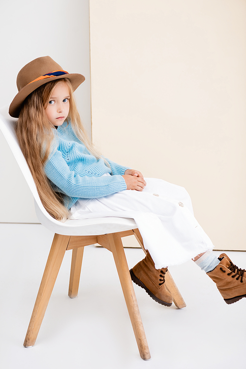 side view of fashionable blonde girl in brown hat and boots, white skirt and blue sweater sitting on chair near beige wall