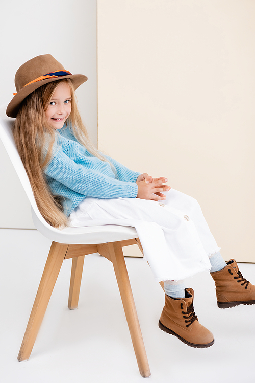 side view of fashionable blonde girl in brown hat and boots, white skirt and blue sweater sitting on chair and smiling near beige wall